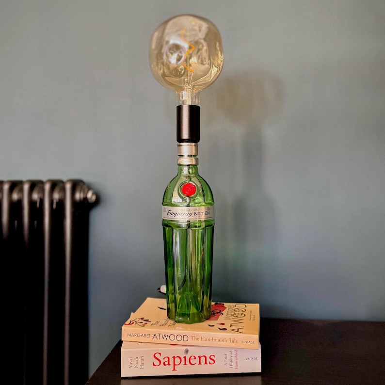 May include: A green glass bottle with a clear glass top and a black light bulb. The bottle is labeled "Tanqueray No. Ten". The bottle is sitting on top of three books: "The Handmaid's Tale" by Margaret Atwood, "A Brief History of Humankind" by Yuval Noah Harari, and "Sapiens" by Yuval Noah Harari.