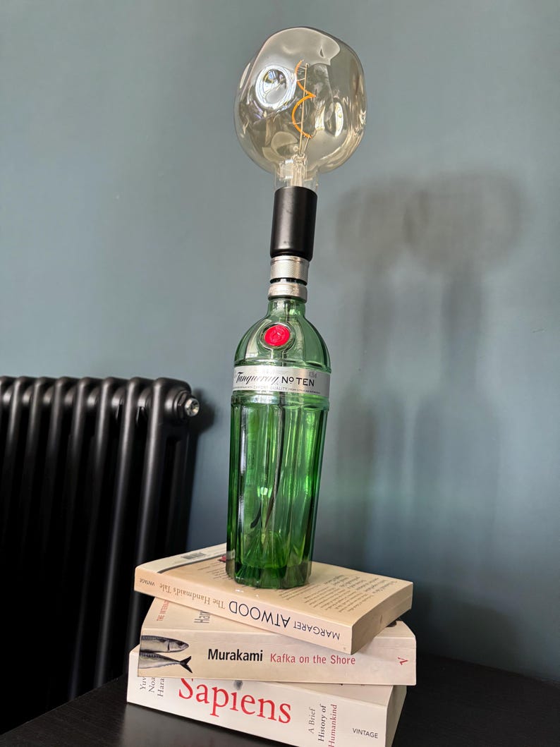 May include: A green glass bottle with a black cap and a clear glass light bulb on top. The bottle is sitting on a stack of three books. The top book has the title "Margaret Atwood The Testaments" on the spine. The middle book has the title "Murakami Kafka on the Shore" on the spine. The bottom book has the title "Sapiens A Brief History of Humankind" on the spine.