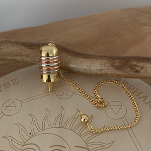 Puede incluir: Un péndulo de oro, plata y cobre con una cadena que cuelga sobre una tabla de madera con un sol y otros símbolos grabados.