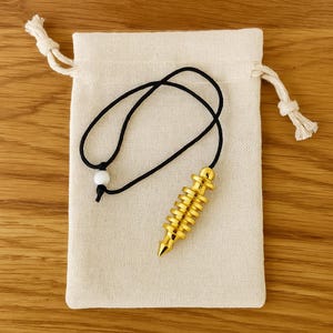 Op de afbeelding: Een goudkleurige pendel met een zwart koord en een witte kraal, op een beige trekkoordzakje. De pendel heeft een conische punt en gestapelde, afgeronde secties. De tas is gemaakt van een natuurlijk gekleurde stof.