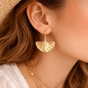 Peut inclure: Boucles d'oreilles en forme de feuille de ginkgo dorée. Les boucles d'oreilles présentent un design texturé et détaillé, avec une fermeture à crochet. Les boucles d'oreilles sont portées sur une oreille, avec un chapeau de paille et des colliers dorés superposés en arrière-plan.