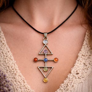 Puede incluir: Un collar con colgante plateado con un diseño geométrico y detalles de piedras preciosas de colores. El colgante cuelga de un cordón negro. El collar presenta un diseño de triángulo y cruz con una variedad de piedras de colores.