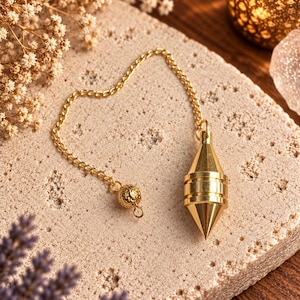 Könnte beinhalten: Ein goldfarbenes Metallpendel mit einer spitz zulaufenden, facettierten Spitze und einem zylindrischen Körper. Das Pendel ist an einer feinen goldfarbenen Kette befestigt, die am Ende des Kettenglieds eine kleine, texturierte, kugelförmige Perle aufweist.