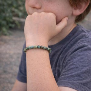 Peut inclure: Un bracelet de perles vertes et marron. Le bracelet est porté sur un poignet, la personne portant un t-shirt gris. Les perles sont petites et rondes. Un bijou simple et naturel.