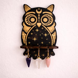 Op de afbeelding: Zwarte houten wanddecoratie in de vorm van een uil met gouden accenten, een plank en drie hangende pendels. De uil heeft hemelse ontwerpen en sterrendetails. De pendels zijn paars, helder en roze.