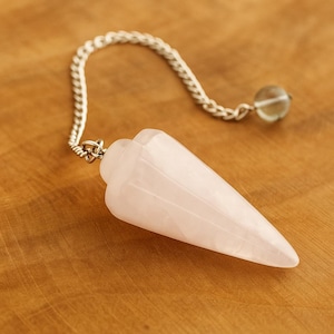 Op de afbeelding: Een rozenkwarts slinger met een zilveren ketting en een kleine, ronde, heldere kristallen kraal. De slinger is lichtroze van kleur en heeft een puntige vorm.
