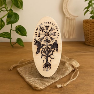 Może przedstawiać: Owalna, jasna drewniana tablica z ciemnym, grawerowanym wzorem. Wzór zawiera centralny symbol, ptaki i drzewo, prawdopodobnie inspirowane mitologią nordycką. Tablica leży na małym worku jutowym.