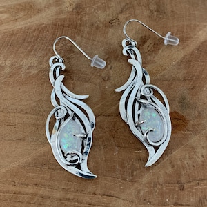 Puede incluir: Un par de pendientes colgantes plateados. Cada pendiente presenta un diseño de hoja ornamentado y estilizado con un centro de piedra blanca iridiscente en forma de pera. Los pendientes tienen un cierre de gancho con un respaldo de plástico transparente.