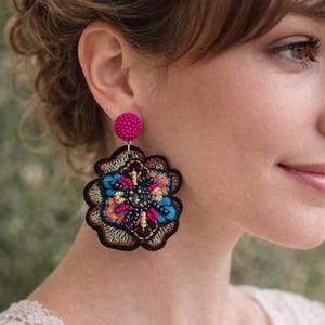 Pode incluir: Close-up de uma mulher usando um par de brincos florais grandes e coloridos. Os brincos apresentam uma base preta com bordados intrincados em tons de azul, laranja, rosa e verde, e um pequeno pino redondo rosa no topo.