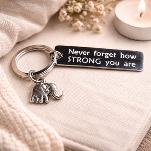 Könnte beinhalten: Silberfarbener Schlüsselanhänger mit einem rechteckigen Anhänger und einem Elefanten-Charm. Auf dem Anhänger steht in weißer Schrift "Never forget how STRONG you are". Der Schlüsselanhänger liegt auf einem hellen Stoff, mit einer brennenden Kerze und Trockenblumen im Hintergrund.