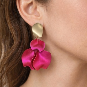 May include: A close-up of a pair of earrings. Each earring features a gold-colored, rectangular top piece and a cluster of three bright pink, petal-shaped dangles. The earrings are a vibrant accessory.