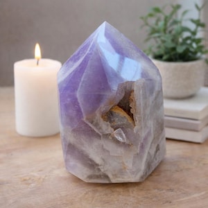 Peut inclure: Une géode de cristal d'améthyste facettée, avec un sommet poli et pointu. Le cristal présente des nuances de violet et de blanc, avec une cavité naturelle révélant un intérieur brun doré. Une bougie blanche allumée est à gauche.