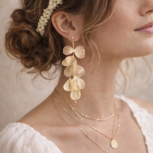 Peut inclure: Boucles d'oreilles dorées en cascade avec un design en forme de pétales superposés. Les boucles d'oreilles sont suspendues à un petit clou et sont associées à de délicats colliers en or. Un accessoire floral pour les cheveux est visible en arrière-plan. Les bijoux sont probablement pour une occasion spéciale.