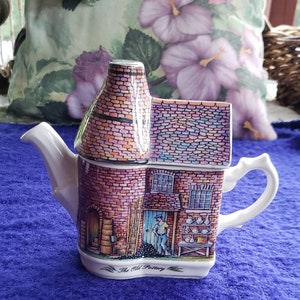 May include: A white ceramic teapot shaped like a brick building with a chimney. The teapot has a colorful brick pattern and a scene of a man working in a pottery shop. The text "The Old Pottery" is printed on the bottom of the teapot.