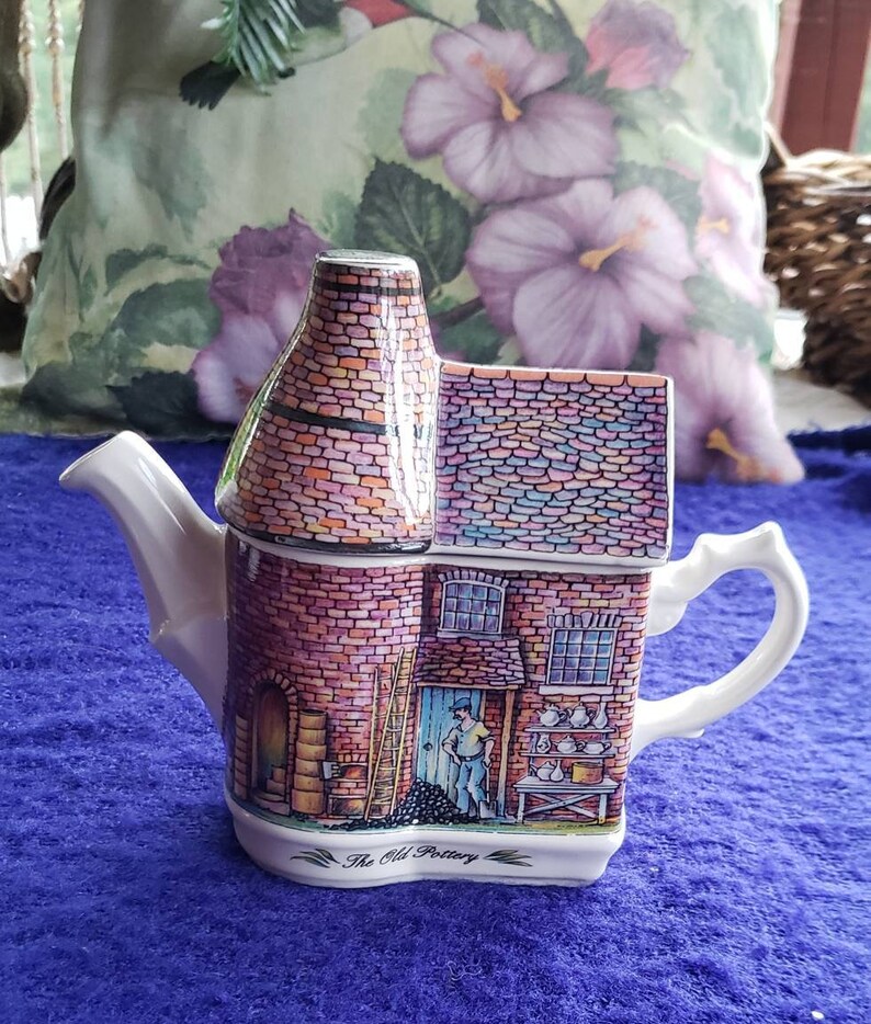 May include: A white ceramic teapot shaped like a brick building with a chimney. The teapot has a colorful brick pattern and a scene of a man working in a pottery shop. The text "The Old Pottery" is printed on the bottom of the teapot.