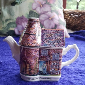 May include: A white ceramic teapot shaped like a brick building with a chimney. The teapot is decorated with a colorful painting of a pottery shop with a sign that reads "The Old Pottery".