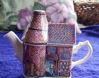 May include: A white ceramic teapot shaped like a brick building with a chimney. The teapot is decorated with a colorful painting of a pottery shop with a sign that reads "The Old Pottery".