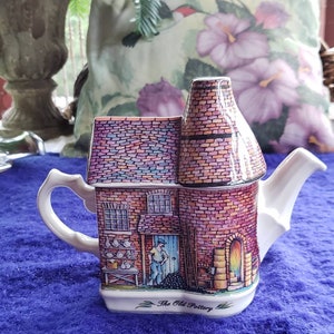 May include: A white ceramic teapot shaped like a brick building with a chimney. The teapot is decorated with a colorful illustration of a pottery shop with a man standing in the doorway. The text "The Old Pottery" is printed on the bottom of the teapot.
