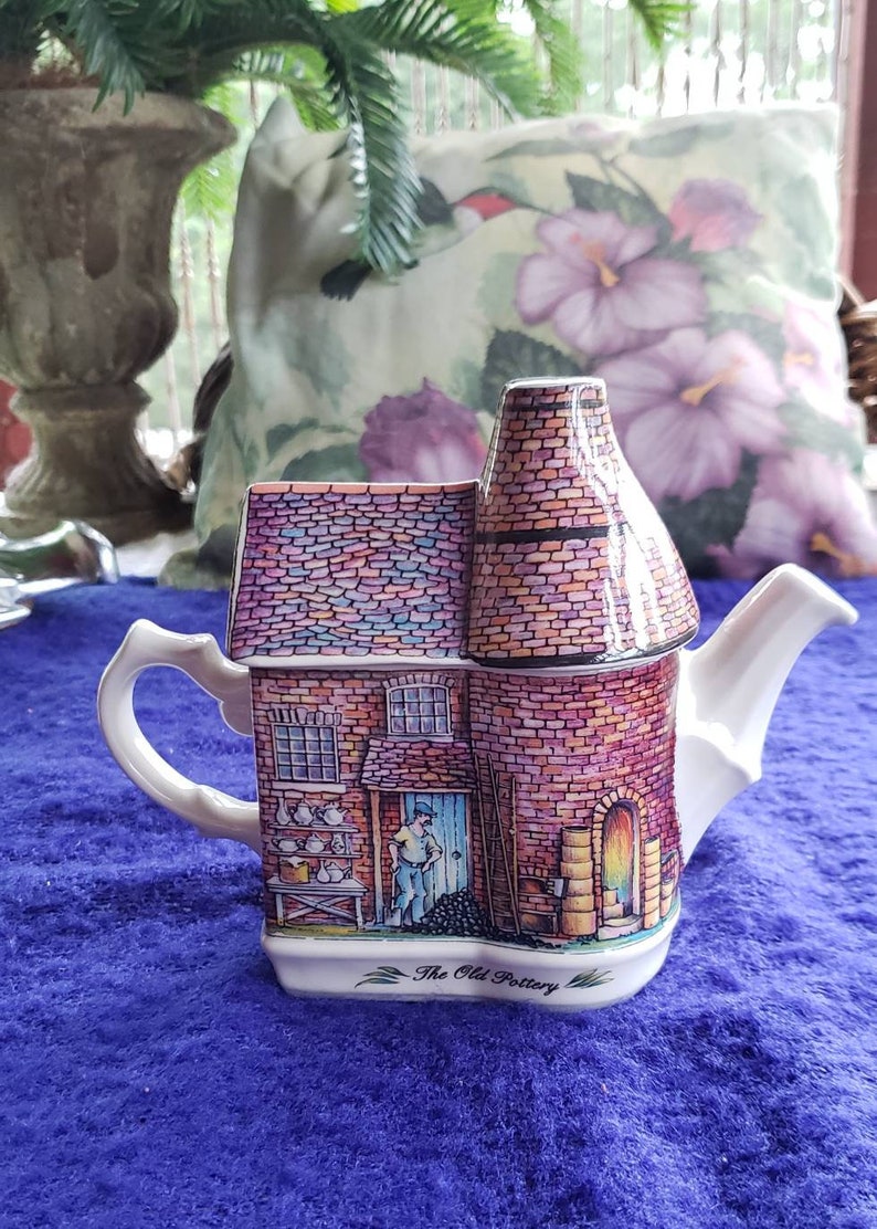 May include: A white ceramic teapot shaped like a brick building with a chimney. The teapot is decorated with a colorful illustration of a pottery shop with a man standing in the doorway. The text "The Old Pottery" is printed on the bottom of the teapot.