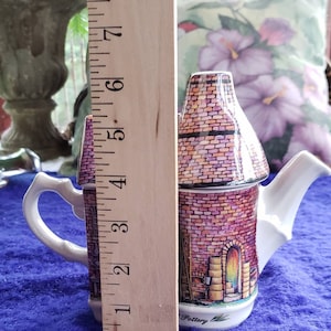 May include: A white ceramic teapot shaped like a brick building with a pink and brown brick pattern. The teapot has a white handle and spout. The teapot is 5 inches tall and is sitting on a blue surface. The teapot has the word "Pottery" written on the bottom.