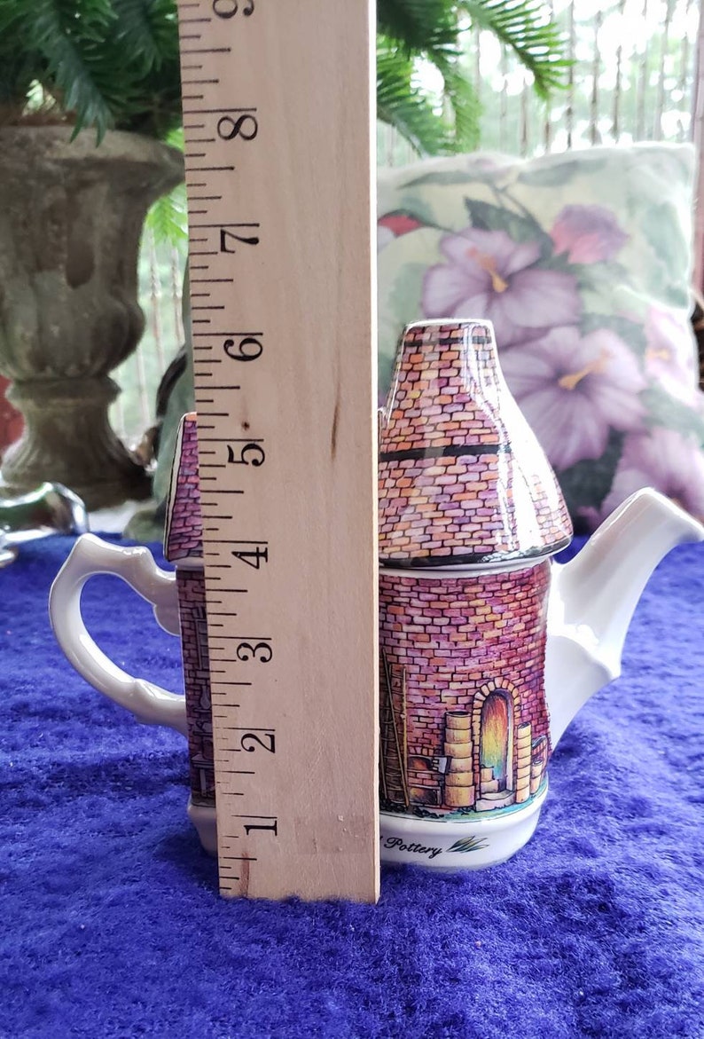 May include: A white ceramic teapot shaped like a brick building with a pink and brown brick pattern. The teapot has a white handle and spout. The teapot is 5 inches tall and is sitting on a blue surface. The teapot has the word "Pottery" written on the bottom.