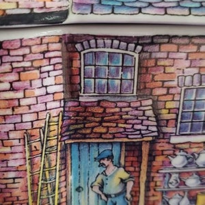 May include: A hand-painted ceramic tile depicting a brick building with a blue door, a ladder, and a man in a blue cap and yellow shirt. The tile has a colorful brick pattern and the text "The Old Pottery" at the bottom.