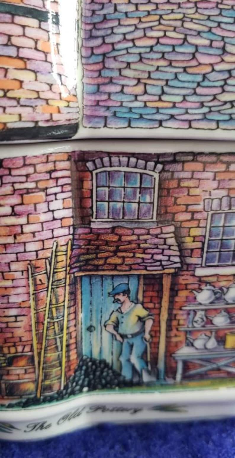 May include: A hand-painted ceramic tile depicting a brick building with a blue door, a ladder, and a man in a blue cap and yellow shirt. The tile has a colorful brick pattern and the text "The Old Pottery" at the bottom.