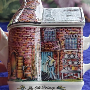 May include: A ceramic teapot shaped like a brick building with a chimney, a ladder, and a man in front of it. The teapot is decorated with a brick pattern, a blue door, and a window. The text "The Old Pottery" is printed on the bottom of the teapot.