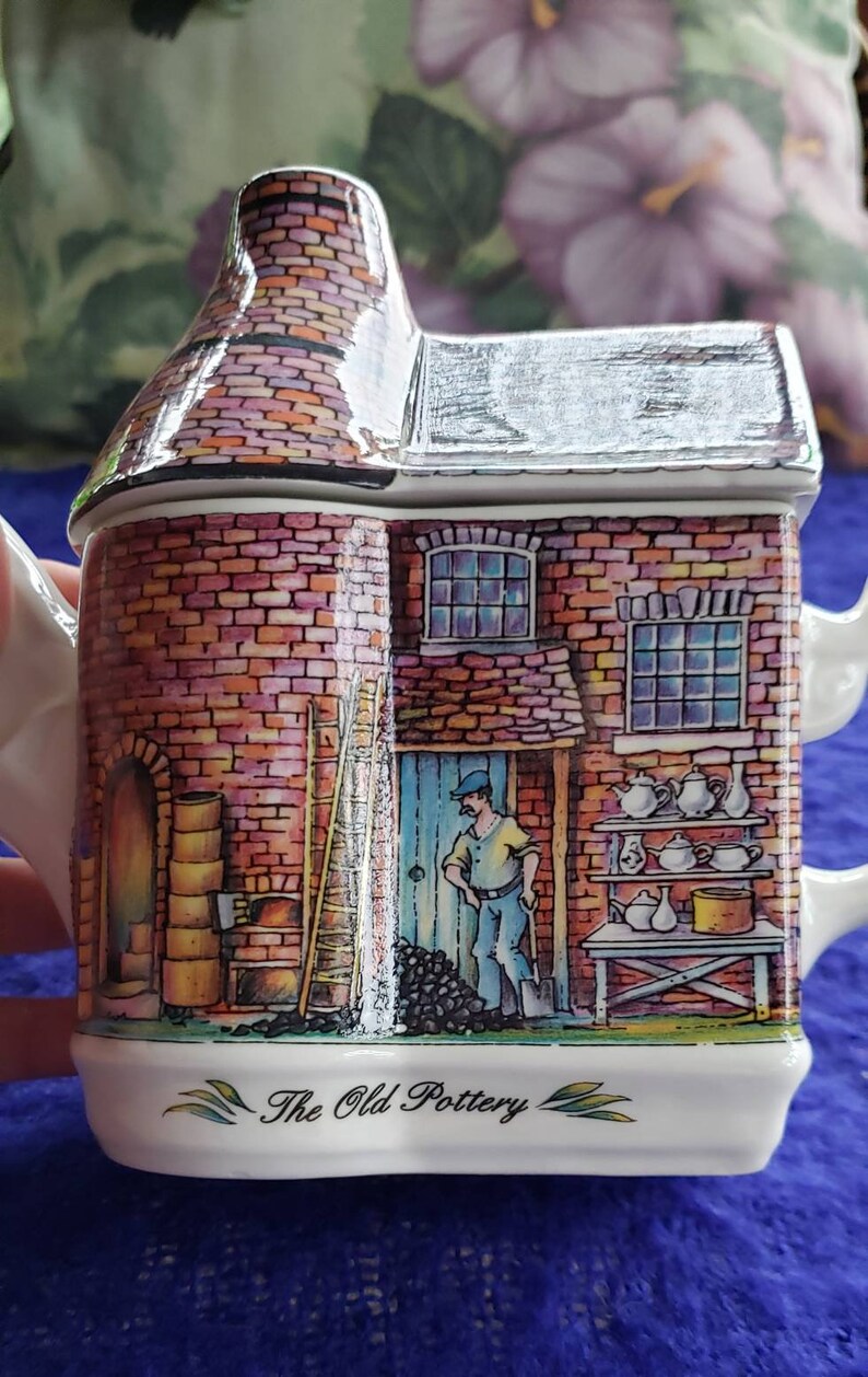 May include: A ceramic teapot shaped like a brick building with a chimney, a ladder, and a man in front of it. The teapot is decorated with a brick pattern, a blue door, and a window. The text "The Old Pottery" is printed on the bottom of the teapot.