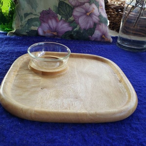 May include: A light brown wooden serving tray with a round glass bowl in the center. The tray has a slightly rounded edge and is made of wood with a natural grain.
