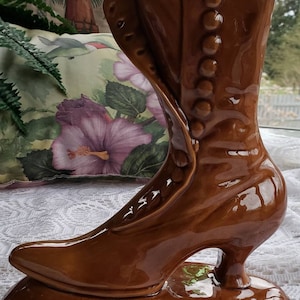 May include: A brown ceramic boot-shaped vase with a high heel and a row of buttons up the side. The vase is glazed and has a shiny finish.