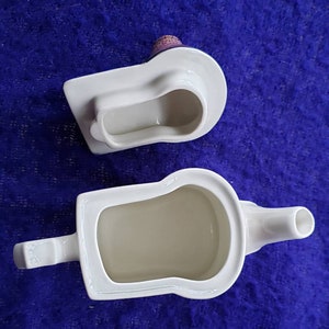 May include: White ceramic teapot with a spout and handle, shaped like a loaf of bread. The teapot is empty and the inside is visible. The lid is separate and is also shaped like a loaf of bread.