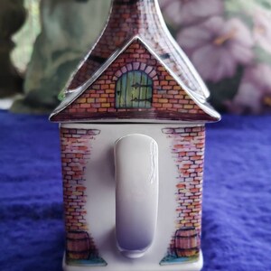 May include: A white ceramic mug shaped like a brick house with a red brick exterior, a green door, and a white handle. The mug has a painted design of a brick wall with a barrel on each side.