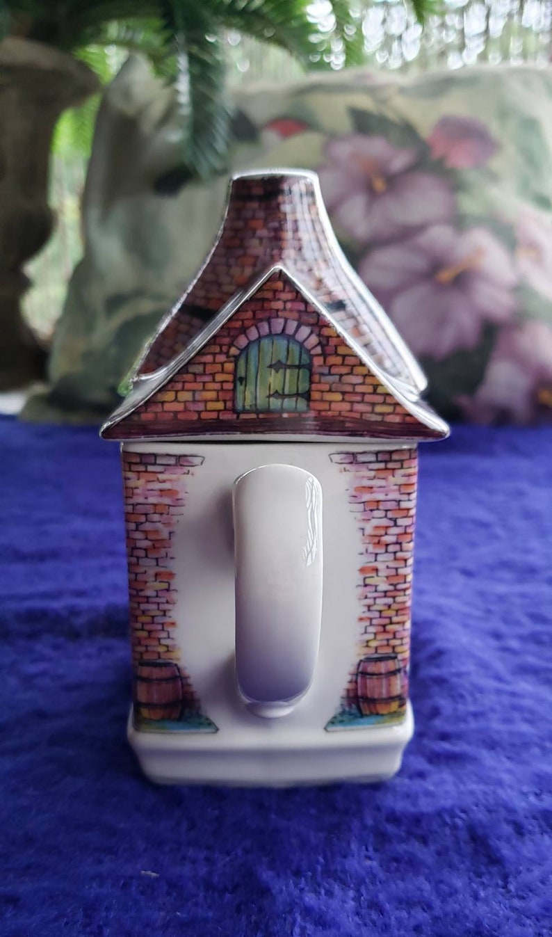 May include: A white ceramic mug shaped like a brick house with a red brick exterior, a green door, and a white handle. The mug has a painted design of a brick wall with a barrel on each side.