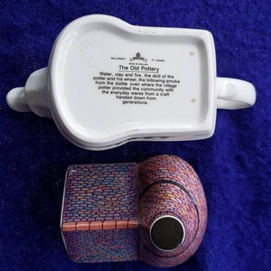 May include: A white ceramic teapot with a colorful brick design. The teapot has a handle and spout. The bottom of the teapot has a label that reads "The Old Pottery" and describes the process of making pottery.