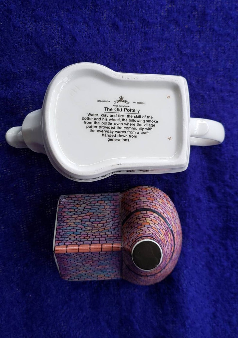 May include: A white ceramic teapot with a colorful brick design. The teapot has a handle and spout. The bottom of the teapot has a label that reads "The Old Pottery" and describes the process of making pottery.