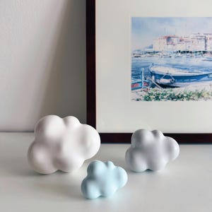Può includere: Tre oggetti decorativi a forma di nuvola, bianchi e azzurri. Sullo sfondo, un dipinto ad acquerello incorniciato di una scena costiera. Le nuvole sono probabilmente in ceramica o porcellana.