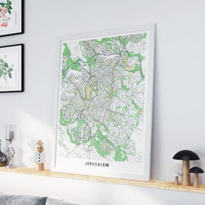 Puede incluir: Un mapa enmarcado de Jerusalén, con un plano detallado de las calles y áreas sombreadas en verde y beige. El mapa está en un marco blanco y se exhibe en una estantería de madera. La palabra "JERUSALEM" está impresa en la parte inferior del mapa.