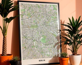 Puede incluir: Un mapa enmarcado de Berlín, Alemania, con borde negro. El mapa presenta un diseño detallado de calles en blanco y negro, con zonas verdes. La palabra "BERLIN" está impresa en negro en la parte inferior. El mapa se muestra sobre una superficie de madera con plantas en macetas.