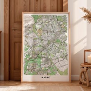 May include: A framed map of Madrid, Spain, with a detailed street layout in shades of green, gray, and beige. The map is encased in a light brown wooden frame and is labeled "MADRID" at the bottom.
