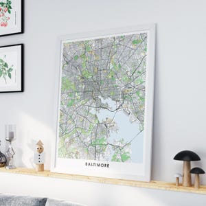 Puede incluir: Un mapa enmarcado de Baltimore, Maryland, en un marco blanco. El mapa muestra las calles, parques y vías fluviales de la ciudad en detalle, con el nombre de la ciudad impreso en la parte inferior. El mapa se exhibe en una estantería de madera.