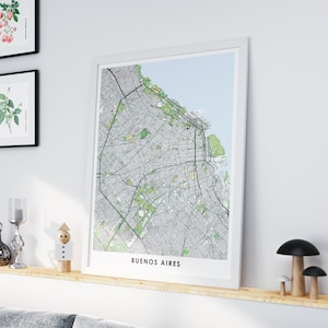 Puede incluir: Una impresión enmarcada de un mapa de Buenos Aires, Argentina, en un marco blanco. El mapa presenta un diseño detallado de las calles con áreas verdes y el nombre de la ciudad en la parte inferior. La impresión se exhibe en una estantería de madera con objetos decorativos.