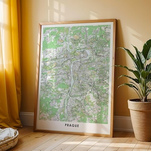 May include: A framed map of Prague, Czech Republic, in shades of green, white, and gray. The map is encased in a light-colored wooden frame and leans against a wall. The word "PRAGUE" is printed in black at the bottom.