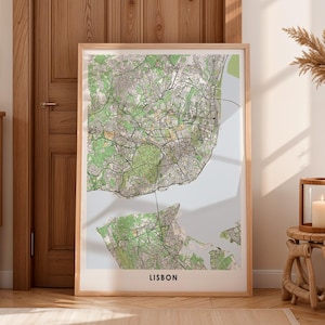 May include: A framed map print of Lisbon, Portugal, featuring a detailed topographical view in green, beige, and gray. The map is set against a white background, with the city's name printed in black at the bottom. The frame is made of light-colored wood.
