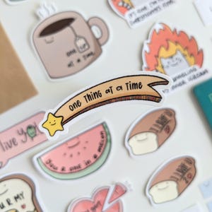 Könnte beinhalten: Eine Sammlung farbenfroher Aufkleber mit verschiedenen Designs. Enthält ein Banner mit dem Text "one thing at a time", einen lächelnden Stern, eine Teetasse, eine Wassermelonen-Scheibe und Cartoon-Brotscheiben. Auf weißem Hintergrund.