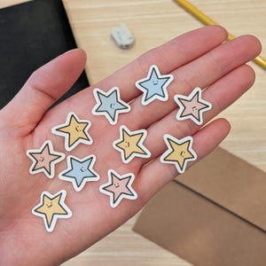May include: A collection of small, die-cut star stickers in pastel shades of yellow, blue, and pink. Each star features a simple, hand-drawn smiling face with two dots for eyes and a curved line for a mouth, outlined in a thin black border.