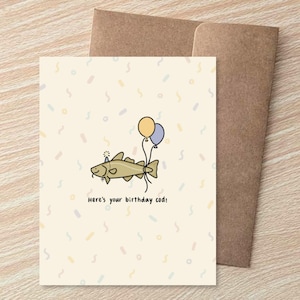 May include: A birthday card with a cartoon codfish holding two balloons. The card has the text "Here's your birthday cod!" and is accompanied by a brown envelope. The background is a light beige with colorful confetti.