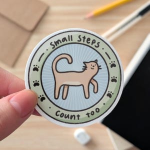 May include: A circular sticker featuring a light brown cat illustration, with the text "Small Steps Count too" and paw prints around the edge. The cat is set against a light blue backdrop with a green border.