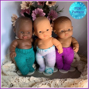 May include: Three baby dolls with varying skin tones, each dressed in knitted trousers in teal, lavender, and purple. The dolls are presented in a decorative setting with flowers and lace trim. A circular "PDF Pattern" graphic is in the upper right corner.