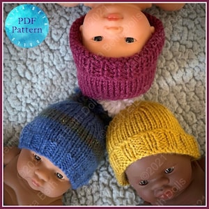 Puede incluir: Tres gorros de punto en diferentes colores: burdeos, azul y amarillo. Los gorros están en la cabeza de tres muñecas.